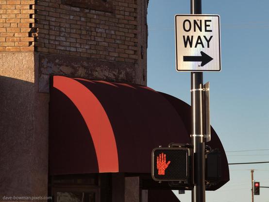 A photograph of a one way sign on Route 66 in Winslow, Arizona, mounted on the same pole as a red-handed stop sign that draws immediate attention. In the background, a brick building with a red awning adds a splash of colour, contributing to the small-town atmosphere and character of this well-known stretch of road.