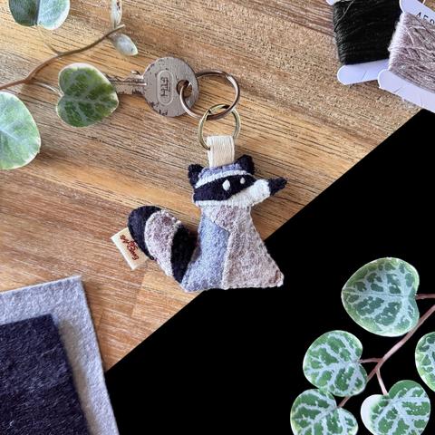 Picture of a handmade felt raccoon keychain on a black and wooden background , a few decorative plants and matching felt and threads around it. The raccoon is facing sideways, but both its white embroidered eyes are on the same side, giving it a silly expression.