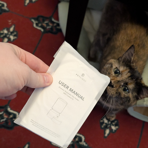 a photo of a tortie cat, Soot. she is sniffing a piece of paper. the piece of paper is being held by the photographer. it is a manual to an automatic pet feeder. 