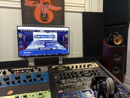 Photo of Total Sonic Media, showing a studio desk filled with analog audio processors, with  B&W Nautilus 802 floor standing  speakers, a pair of Beyer Dynamic headphones - with a large monitor displaying the TSM website on the wall, along with a bright orange & brown abstract painting of a guitar emitting sound waves, and various acoustic treatments.