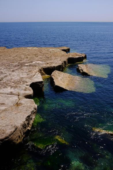 A view out to sea showing a flattish area of stone, once the quay. Two parts of the edge have fallen off into the sea after being undermined by the waves. The sea and the sky are blue.