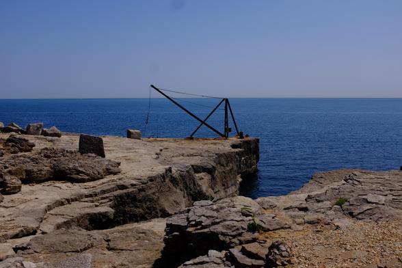 Looking out from the land to the sea, at the edge is an iron hoist silhouetted against the blue sea.In the foreground is a cliff shaped as a quay in the past but eroded away in places.