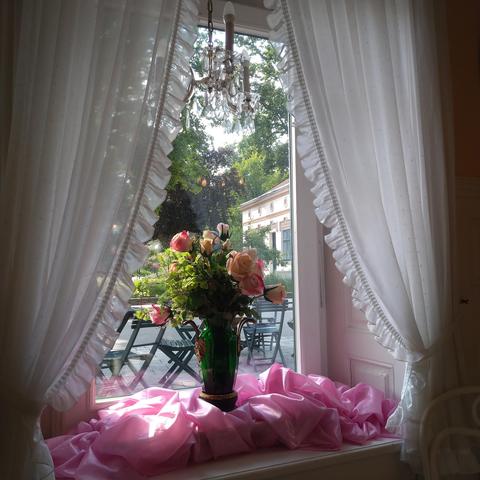 Ein Fenster gerahmt mit Rüschen Vorhängen, auf dem Fensterbrett steht eine Blumenvase zwischen rosa Tüll