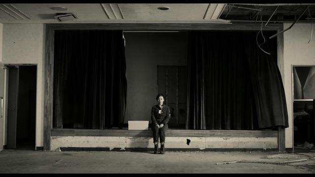 A black-and-white photo of a woman sitting alone on a stage in an empty, abandoned space. The stage has black curtains on either side and a white box in front of her. The room appears neglected, with visible wear and tear. 