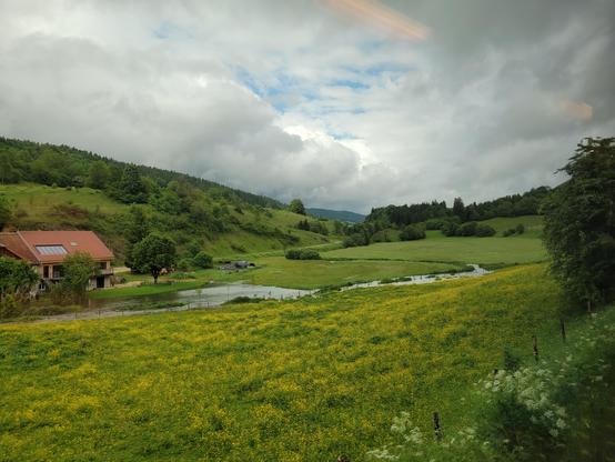 View from the window: valley with green grass and a touch of yellow flowers, under rainy clouds and a touch of blue sky.