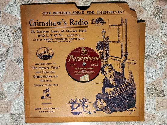 A 78rpm gramophone record in a cover. 

The cover is brown card with black lettering - 
Our records speak for themselves!
Grimshaw's Radio
25, Rushton Street & Market Hall
Bolton. Telephone Bolton 2439.
And as Radio Corner, Ormskirk
Telephone Ormskirk 14.
Accredited Agent for His Masters Voice and Columbia Gramophones and Records.
Complete Stocks Held.
Easy Payments Arranged. 

There is a line drawing over approx 1/3 of of the cover, showing the edge of a building, with a drainpipe, and a streetlight in the background. The foreground is an older male sat on the floor, his back against the building, playing an accordion and operating two small dancing puppets on a line attached to his shoe.

The record by Parlophone has a burgundy label with text in gold. The Song is The Warblers Serenade, played by Frank Westfields's Orchestra, Prince of Wales Playhouse, Lewisham with Whistling by James Bendon. It also has and orange and white 1d value stamp.