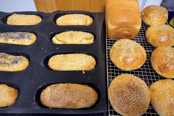 Freshly baked bread: hot dog buns in a silicone mold and a loaf of sandwich bread cooling on a cooling rack next to sesame seed covered hamburger buns.