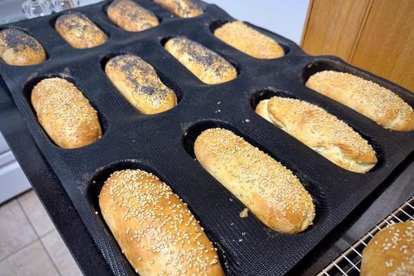 Six sesame seed topped hot dog buns and six poppy seed hot dog buns in a silicone mold freshly out of the oven.