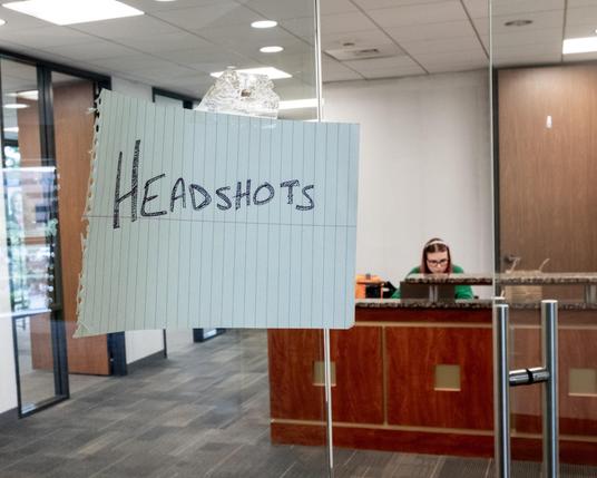 Paper sign with handwritten word “HEADSHOTS” taped to the glass door of an office. In the background, a person sits behind a desk, looking down.