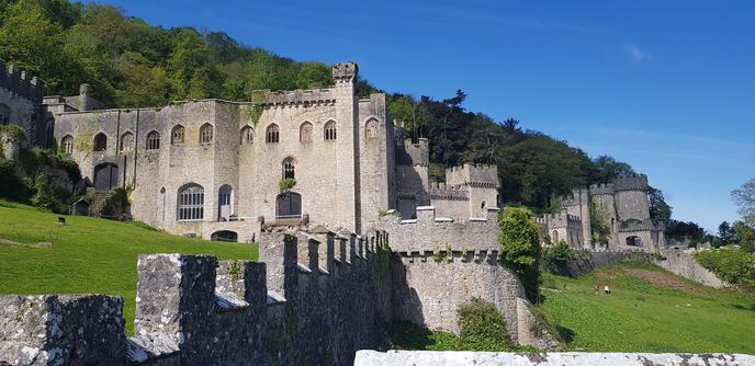 A historic stone castle with multiple towers and battlements, set against a lush green hillside under a clear blue sky. The structure features arched windows and a fortified wall, with greenery climbing parts of the stonework. A grassy foreground and a few people walking add to the scenic view.
