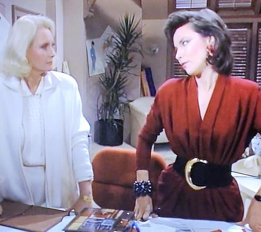 Two women standing side-by-side glaring at each other. Obviously, soap opera enemies having words. Woman on right wearing a burgundy-colored V-neck dress with a MASSIVELY WIDE black belt with gold buckle.