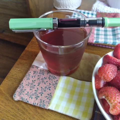 Photo of a TWSBI Eco balanced across a glass of ice tea. The body of the pen is clear resin with a dusty green cap and end. The ink inside is a dark purple brown. A bowl of strawberries to the side.