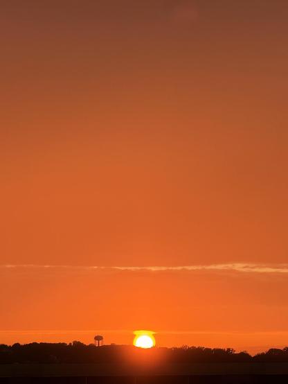 The setting sun with an orange-red sky above it. There’s some silhouetted land and a water tower closer to us than the sun