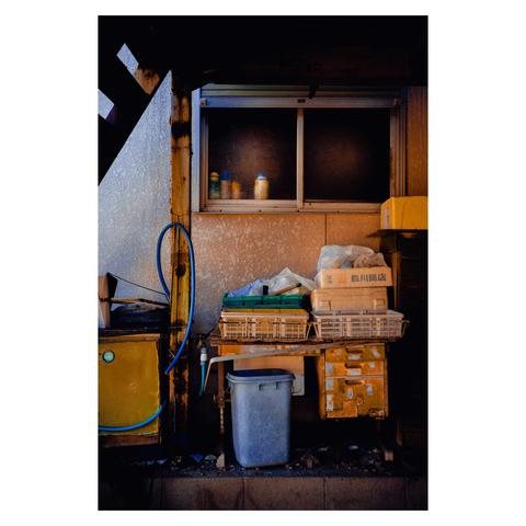There’s a weathered metal sink unit to the left with a blue hose coiled against a rusty yellow surface. In the center, a cluttered wooden table holds plastic baskets, styrofoam boxes, and plastic bags—objects likely used for deliveries or outdoor prep work. Beneath it sits a slightly grimy trash bin. To the right, a small drawer unit, once white or yellow, shows heavy signs of age and rust. Above it all, a frosted glass window glows softly from the inside, with colorful spray cans or bottles lined up on the sill, catching the light.