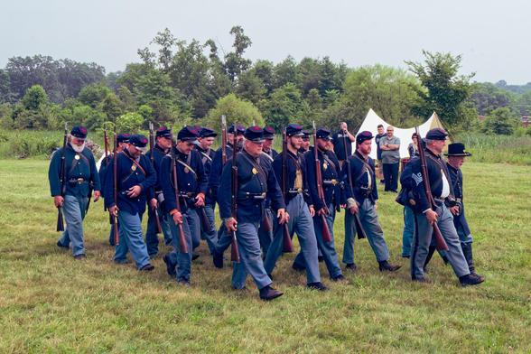 About twenty soldiers march from the rear left towards the front right on closely cut grass with white tent and row of bushes behind them.