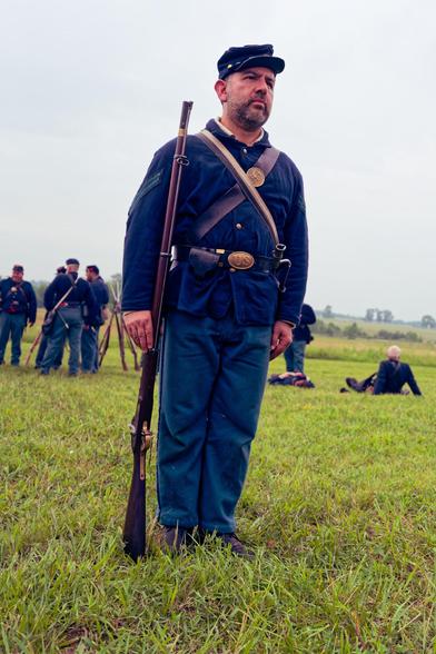 Man with short beard wears a blue jacket and blue hat and blue pants and holds a long black powder rifle pointing straight up that reaches almost to his ear while a few more soldiers are at ease behind him