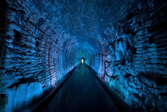 Inside the historic Brockville Railway Tunnel in Brockville, Ontario, Canada’s oldest railway tunnel. The image shows a long, arched tunnel made of rough stone, dramatically lit with blue and purple lights. The tunnel appears empty and stretches straight ahead, with the walls and ceiling glowing in cool colors. At the far end, there is a small, bright opening filled with white light, creating a strong sense of depth and perspective. The overall mood is mysterious and peaceful, with the tunnel’s textures and colors standing out vividly.
