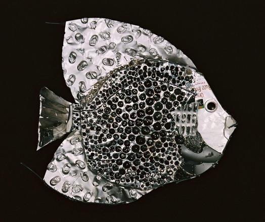 An assemblage of a fish made from blister packs, feathers, toothbrush, wire, coffee bag, buttons, snaps, beads, acoustic tiling, and textured plastic debris, forming a silvery-gray angelfish against a black background.