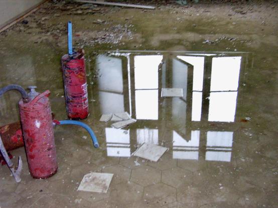 In einer Pfütze auf dem Boden spiegelt sich ein offenes Sprossenfenster. Links stehen zwei Feuerlöscher. Der Boden ist blanker Estrich.