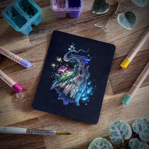 A photo of a small black paper painting placed on a wooden table and surrounded by coloured pencils, a paint brush, paint tubs, and leaves for decoration. The painting features a seemingly hovering twisty log covered in moss and various colourful, glowing mushrooms. 