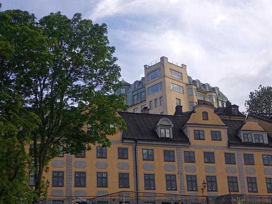 Orange four storey building with many windows, behind it a light yellow building built on higher ground with large studio windows on the two top floors on the corner. The crown of a large tree to the left in the picture.