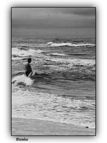 Black-and-white photo of a man standing in the sea, holding an inflatable swim ring. The waves reach up to his thighs — they are choppy and foamy. The man is turned slightly to the side, and his posture suggests he has just entered the water or is about to submerge himself. In the background stretches a dynamic sea and a blurred, cloudy sky. The shot has a raw, documentary quality and captures a moment of spontaneous bathing in the cold, turbulent sea.