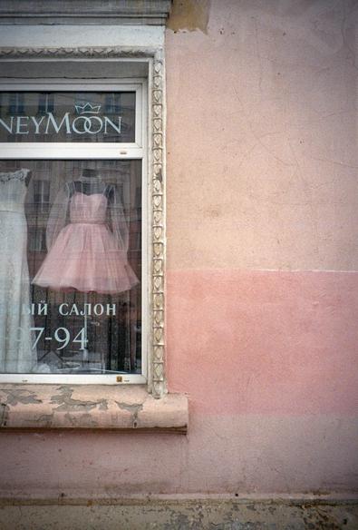 The photo shows the wall of an old pink house and part of a window. Two wedding dresses are visible in the window: a pink one and a white one.