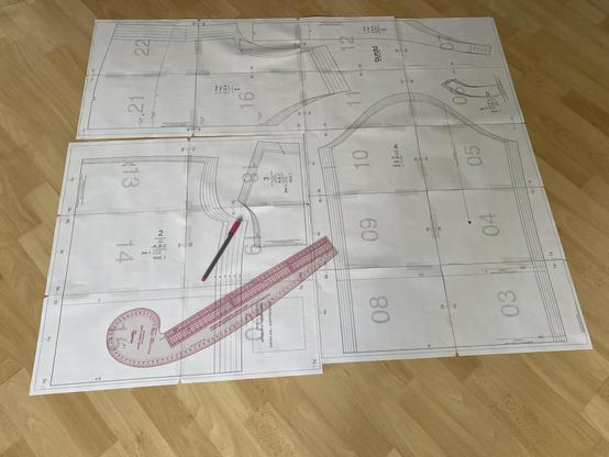 Ein großer, weißer Bogen Papier auf dem Boden liegend (Holzoptik) bedruckt mit einem Schnittmuster verschiedener Teile einer Bluse. Auf dem Papier liegt ein pinkfarbener Stift und ein 50 cm langes, gebogenes Lineal. 