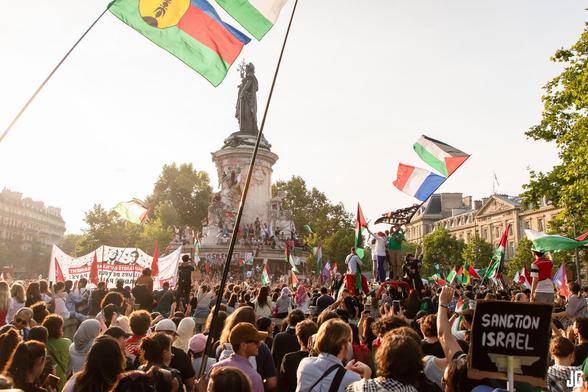 Une large foule se rassemble autour de la statue centrale de la place. De nombreux drapeaux sont brandis, dont un drapeau français et un drapeau palestinien attachés ensemble. Au premier plan, une pancarte portant l’inscription « Sanction Israel » est bien visible.