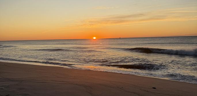 The sun begins to rise over the horizon of the sea on a clear, calm morning.  Small waves are on the sea's surface.  