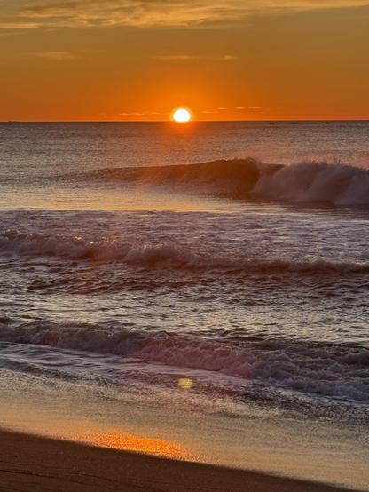 The sun begins to rise over the horizon of the sea on a clear, calm morning.  Waves are breaking.  Colors are intensified reds and oranges
