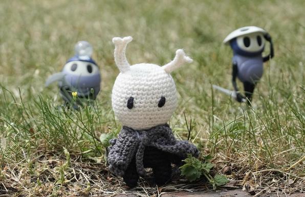 A crocheted Holliw Knight character with a white head and gray cloak stands in the foreground of a grassy area, flanked by two smaller figures in the background, one with a blue hat and the other with a sword.