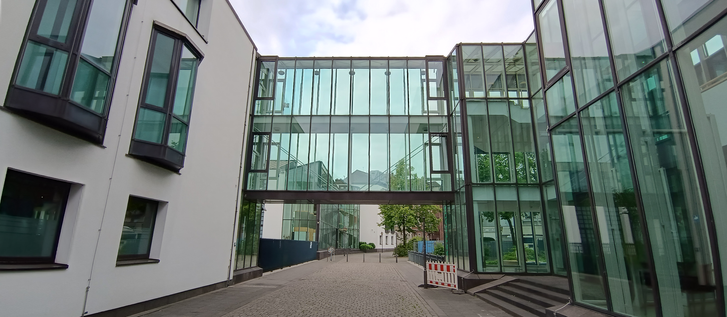 Die Abbildung zeigt einen modernen Gebäudekomplex, der durch eine verglaste Brücke verbunden ist. Links ist ein weißes Gebäude mit dunklen Fensterrahmen zu sehen, während rechts ein weiteres Gebäude mit einer überwiegend gläsernen Fassade steht, das in einem türkisfarbenen Ton schimmert. Die gläserne Brückenkonstruktion, die die beiden Gebäude miteinander verbindet, spiegelt den Himmel und die Umgebung wider. Unterhalb der Brücke führt ein gepflasterter Weg durch den Komplex, der in der Ferne Bäume und weitere Gebäude erahnen lässt. Die Architektur wirkt funktional und hell, mit einem Schwerpunkt auf Transparenz und modernen Materialien.