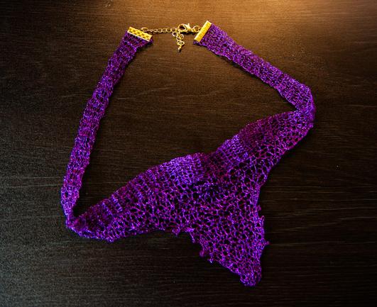 Handmade crochet necklace in vivid purple, shaped like an organic root or branching structure. The asymmetrical design evokes natural forms, with fine openwork resembling tendrils or veins, and is finished with a gold clasp and chain extender. Set against a dark wooden background.