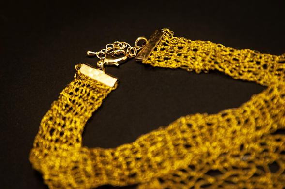 A handmade crochet necklace crafted from shimmering golden thread. The piece features a central spiral motif resembling a blooming flower or radiant sunburst, attached to a wide mesh band and finished with a gold clasp and chain. Displayed on a dark background that enhances the metallic glow.