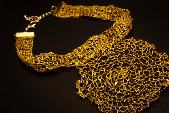 A handmade crochet necklace crafted from shimmering golden thread. The piece features a central spiral motif resembling a blooming flower or radiant sunburst, attached to a wide mesh band and finished with a gold clasp and chain. Displayed on a dark background that enhances the metallic glow.