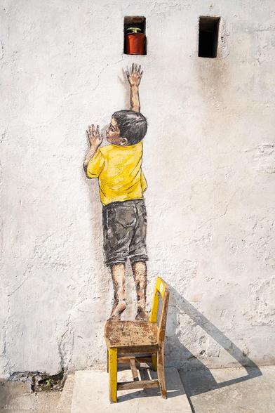 A photograph of a mural showing a young boy standing on a real wooden chair, reaching up toward a painted window on a worn, textured wall in George Town, Penang, Malaysia. The artwork is part of Ernest Zacharevic’s street art series, blending painted elements with physical objects to create a playful, interactive effect that merges art with its surroundings.