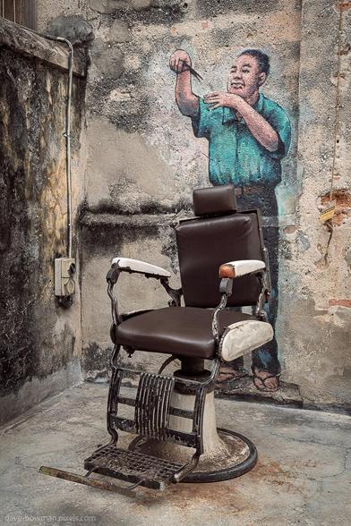 A photograph of an interactive wall mural in the Kwai Chai Hong area of Kuala Lumpur, Malaysia. The artwork includes a real vintage barber’s chair placed in front of a painted scene showing a barber mid-trim, creating the illusion that the seated visitor is having their hair cut. The realistic alignment between the physical chair and the mural adds a playful and immersive element to the scene.