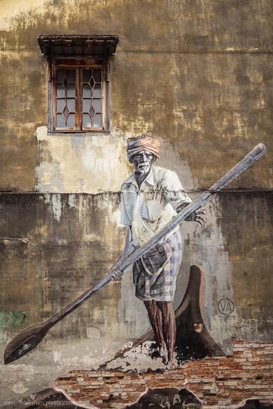 A photograph of the Indian Boatman wall mural in Penang, Malaysia, showing a painted figure rowing a wooden boat across a textured wall. The mural is detailed and lifelike, with the boatman’s posture suggesting gentle movement through water. The rough, weathered surface of the wall becomes part of the scene, blending with the artwork to create a sense of quiet motion and a link to everyday local life.