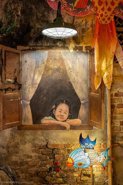 A photograph of a wall mural in the Kwai Chai Hong area of Kuala Lumpur, Malaysia, showing a painted young girl leaning through an open window. The scene is lit by an overhead lamp, creating a seamless blend between the artwork and its surroundings. In front of the mural, a brightly coloured fish with an internal light, added as part of the Chinese New Year decorations, provides a vivid contrast against the more muted tones of the wall.