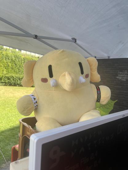 Das Plushtodon sitzt mit Eintrittsbändchen an beiden Armen auf einem Getränkekiste
