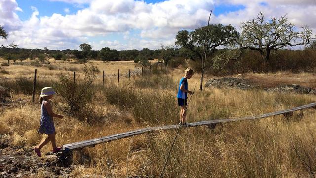 Two children walk along a narrow wooden footbridge through dry grassland at Bubulcus & Bolotas. One wears a sunhat and dress, the other carries a stick. Sparse trees and a wire fence stretch across the warm, maditerranean landscape under a partly cloudy sky.