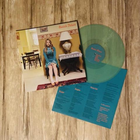 Das Cover des Albums "Basia's Palace" zeigt eine Frau, die in einem Haus sitzt, mit einer grünen Schallplatte daneben. Die Szene umfasst eine Lampe, einen Stuhl und einen dekorativen Teppich. Die Verpackung enthält auch einen türkisfarbenen Einleger mit den Songtiteln.