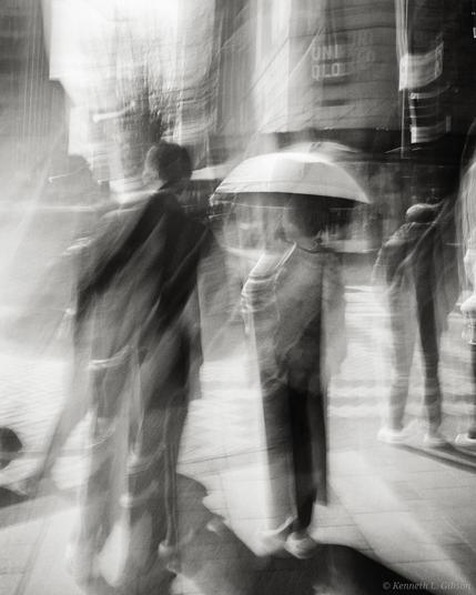 Black and white ICM photo of people on a street corner.  One has an umbrella.