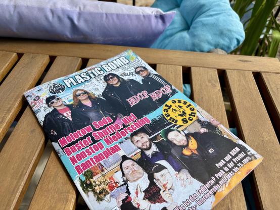 A magazine titled "Plastic Bomb" featuring a colorful cover with images of a group of people in punk attire, including leather jackets and sunglasses. The magazine features articles about various punk bands. The setting includes a wooden table and cushions in the background.