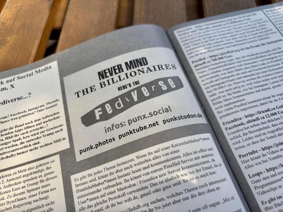 An open pamphlet on a wooden surface featuring an advertisement titled "Never Mind the Billionaires, Here's the Fediverse." The ad provides information about various alternative social media platforms, with website links listed. The text is primarily in German, discussing the Fediverse.