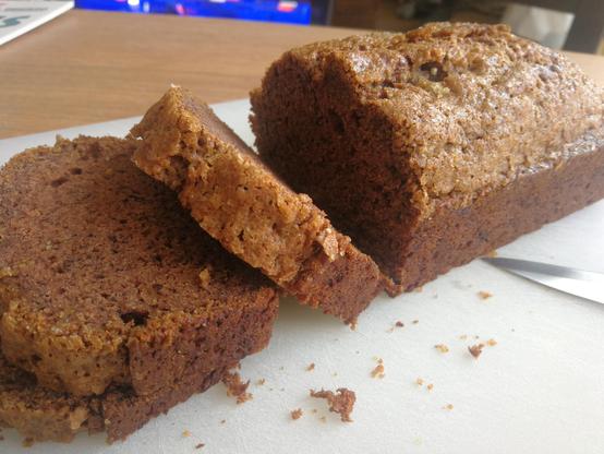 Bruine bananencake op een snijplank, met enkele afgesneden plakken. De cake is mooi gerezen, en er zit een suikerlaagje op.