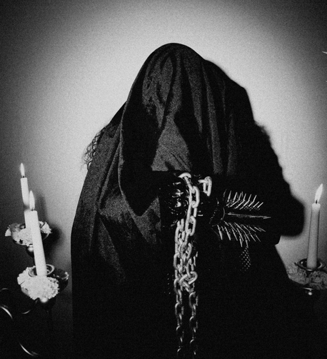 photo of a hooded figure, holding chains, wearing a spiked gauntlet, flanked by candles.
