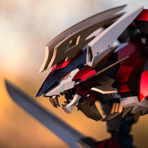 Close-up of a Hayate Liger Zoid, a mechanical lion with sleek red, gold, and silver armor plating. Its angular head is detailed with sharp fangs, vents, and blade-like fins, giving it a fierce, futuristic appearance. A sword is visible in the background, adding to the dynamic composition.