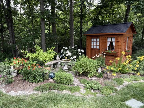 Garden with flowers and a cute garden shed in the background.  There is a concrete bench, bird bath and statue.  